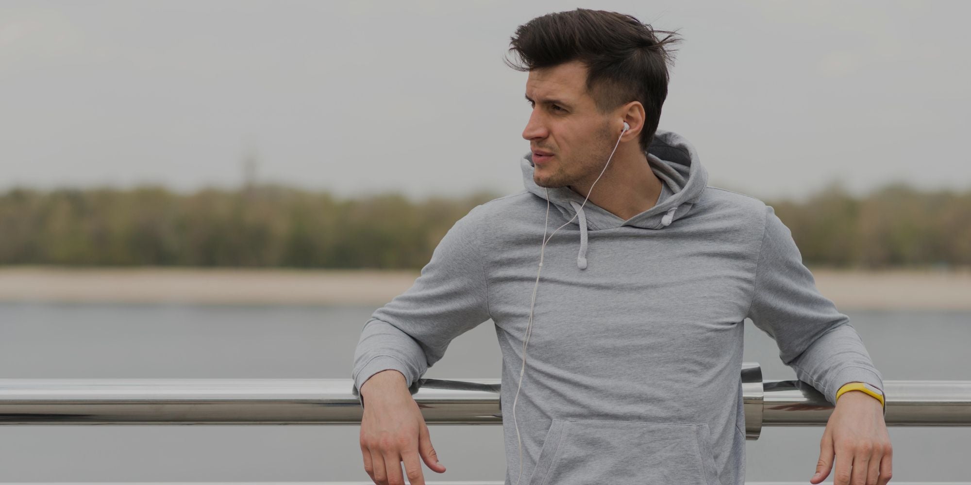 A man in a gray hoodie leaning on a railing outdoors, wearing white wired earphones, with a blurred lake and trees in the background.