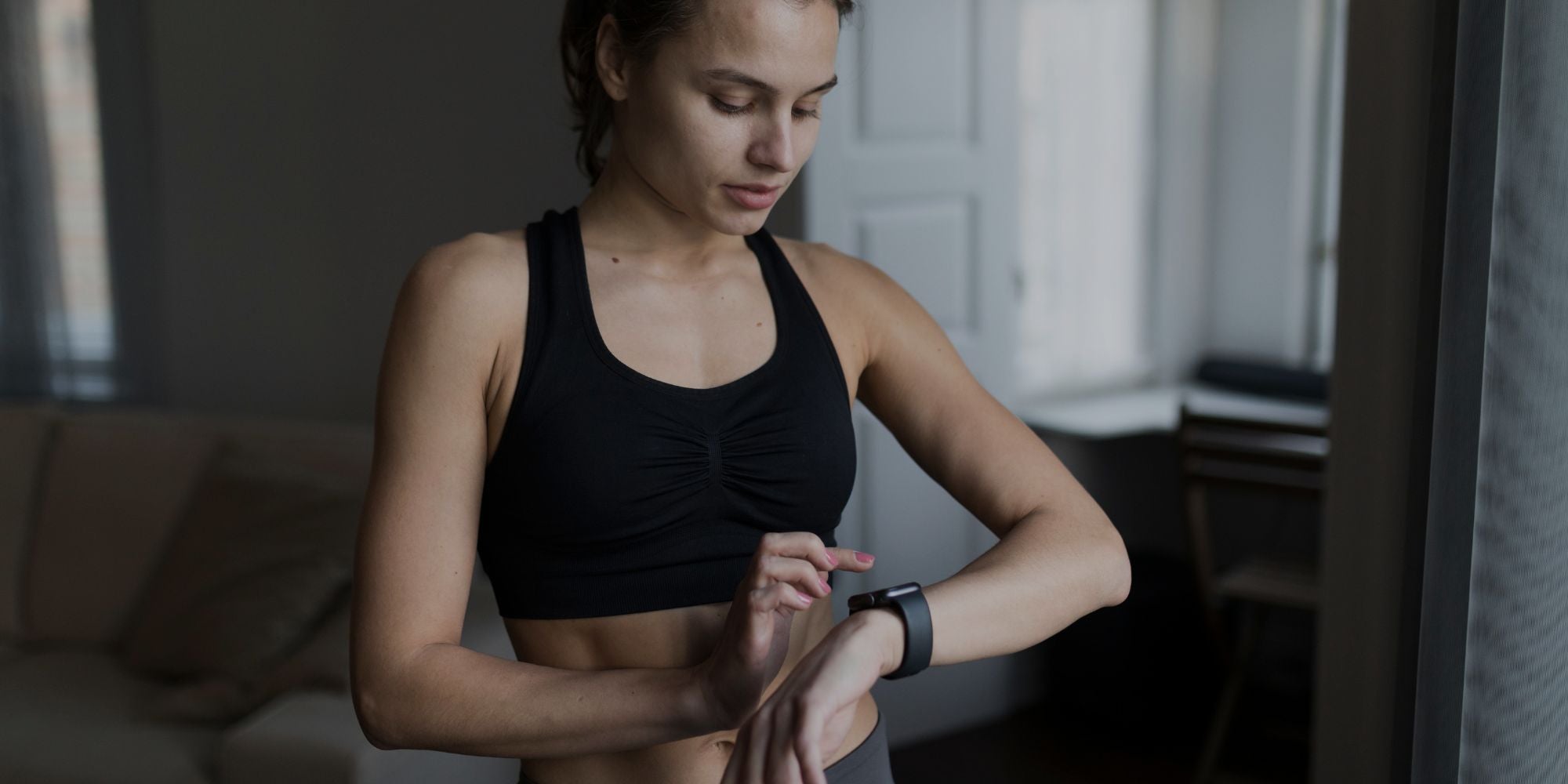 A woman in a black sports bra checks the screen of a black smartwatch in a dimly lit indoor room.